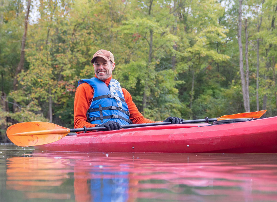 A man in an orange shirt and a blue vest sits in a red kayak on a river with trees behind him.