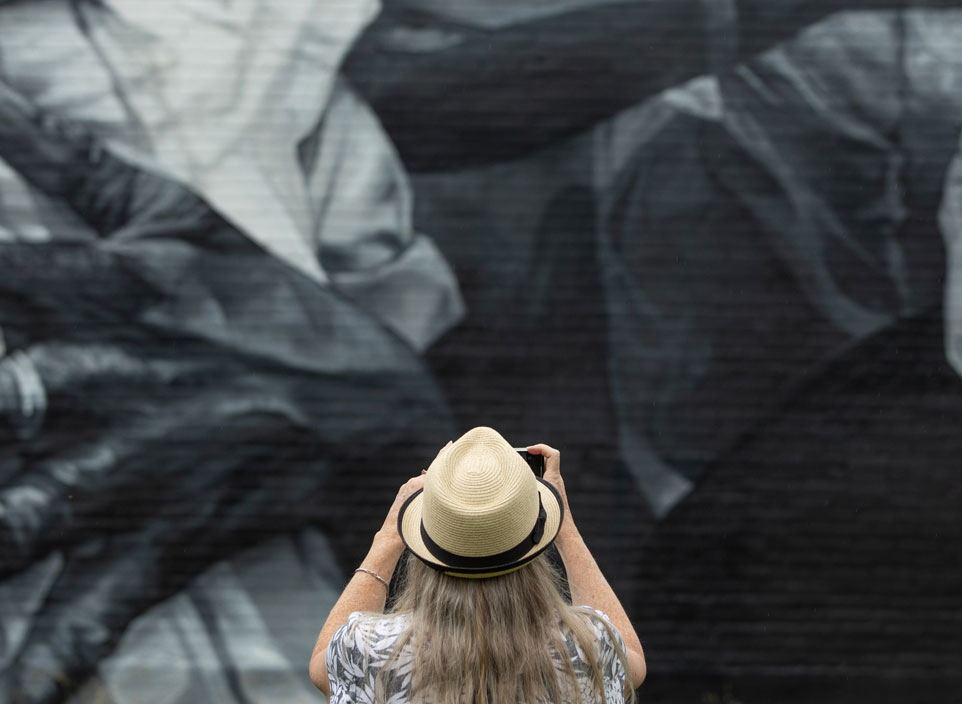 A woman in a straw hat takes a photo of a black and white mural.